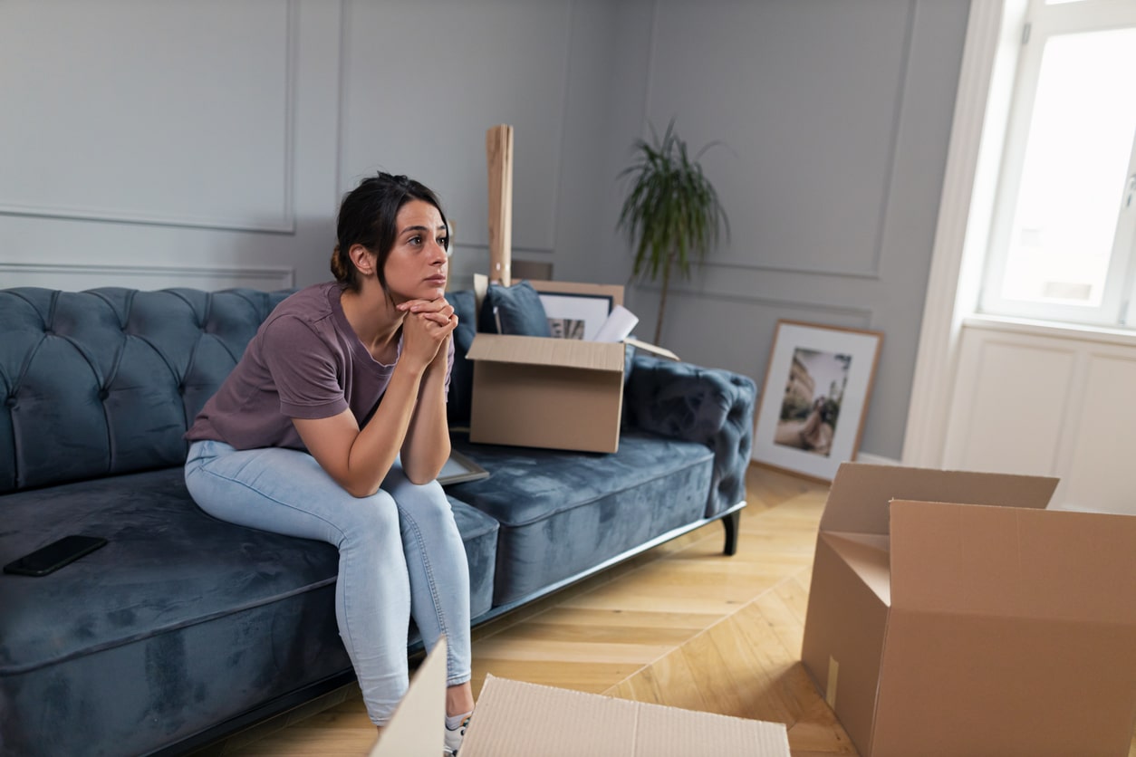 A woman sits on a sofa surrounded by moving boxes in a room contemplating moving after a natural disaster