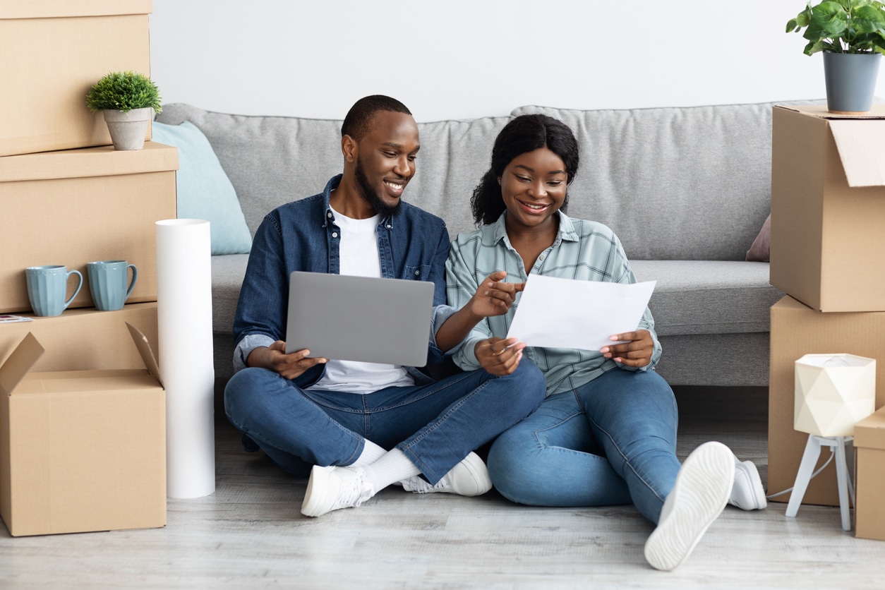 A couple sits on the floor surrounded by moving boxes, smiling while looking at documents and a laptop