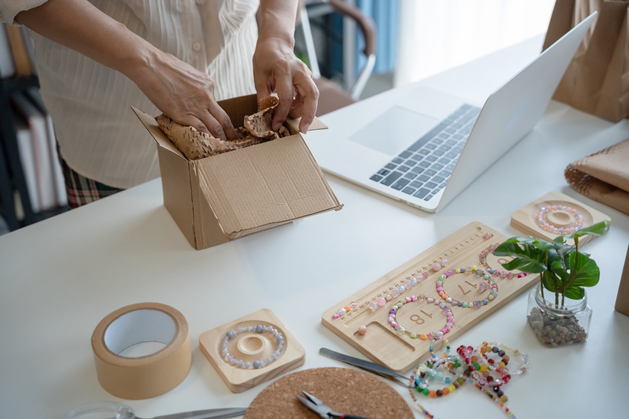 Hands packing a small shipping box with crinkle paper, next to bracelets, jewelry supplies and a laptop