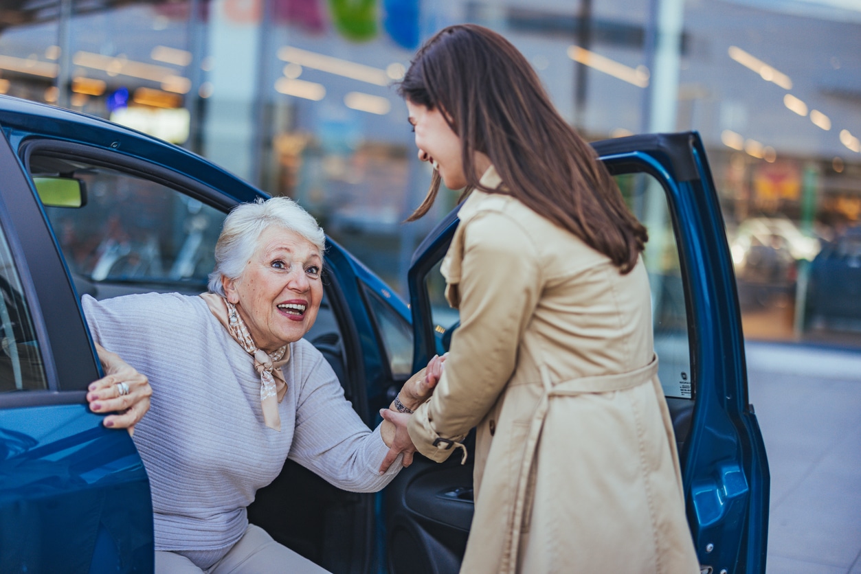 Happy Elderly Woman Assisted by Caring Young Woman Outdoors A younger woman helps her smiling elderly mother out of a blue car as she assists her with moving to another state