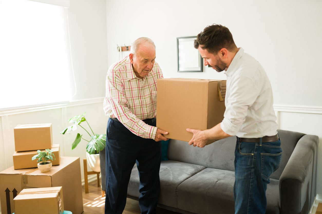 A younger man helping his elderly father carry a large moving box as he moves to a new home out of state