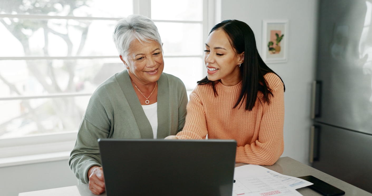 Kitchen, mother and woman with laptop for finance, track expenses and check budget goals. Discussion, senior mom and daughter with tax review for retirement, handle savings and mortgage at home A younger woman and an elderly woman smile while looking together at elderly parent moving options on a laptop