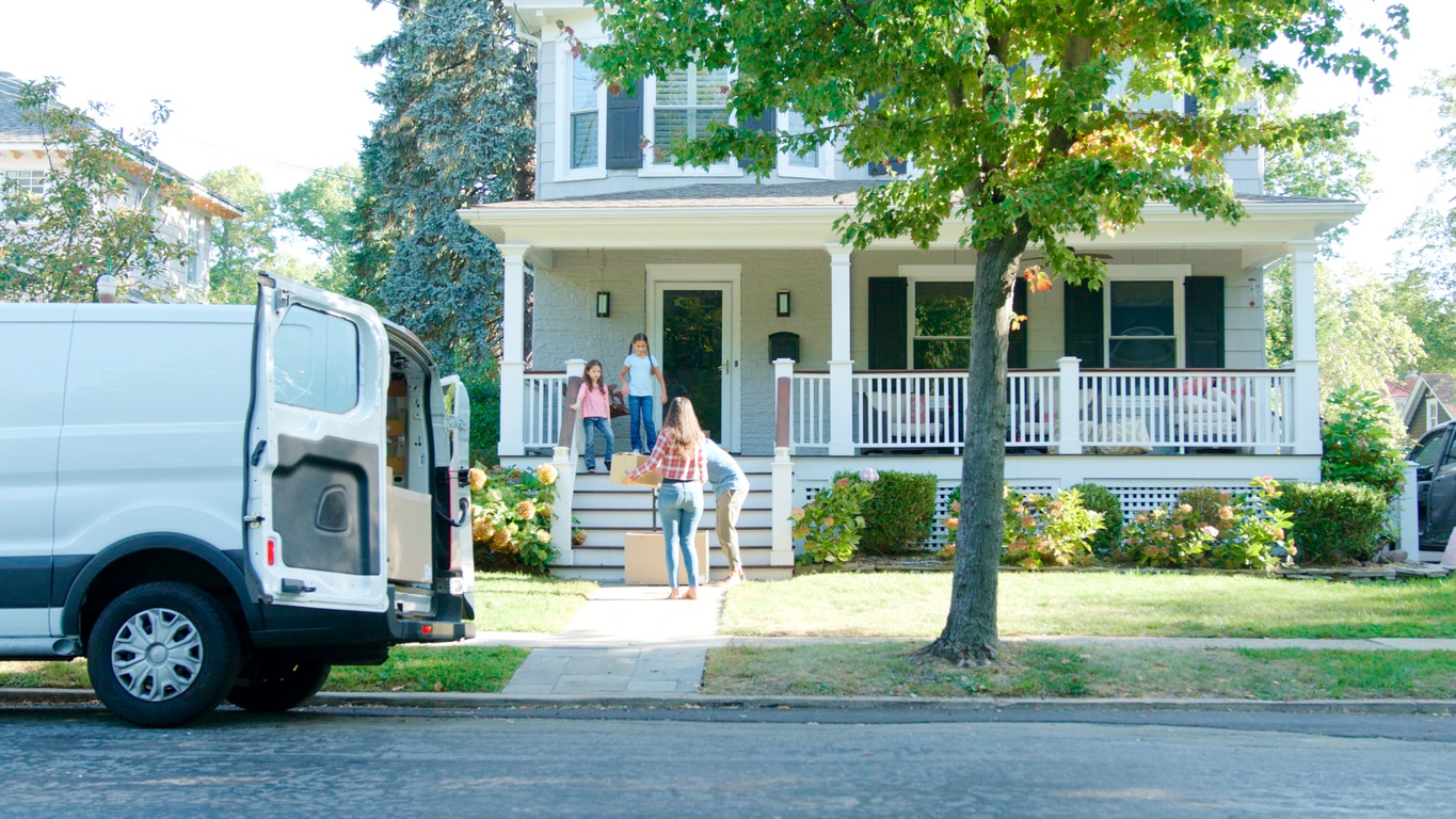 A family unloads moving boxes from a moving van into their new two-story home on a sunny day