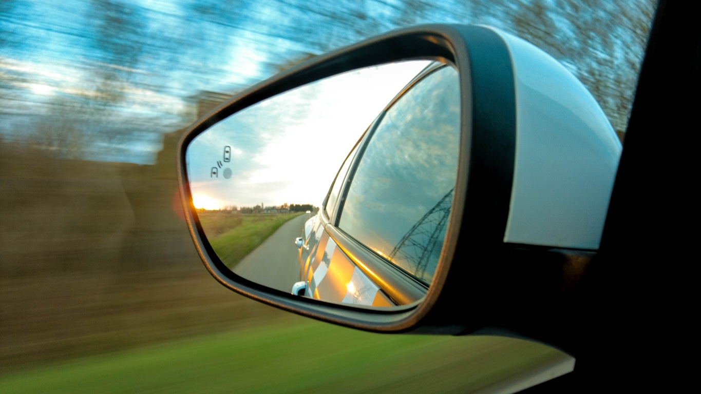 A close-up of a car's side-view mirror reflecting a sunset over a road, with motion blur in the background
