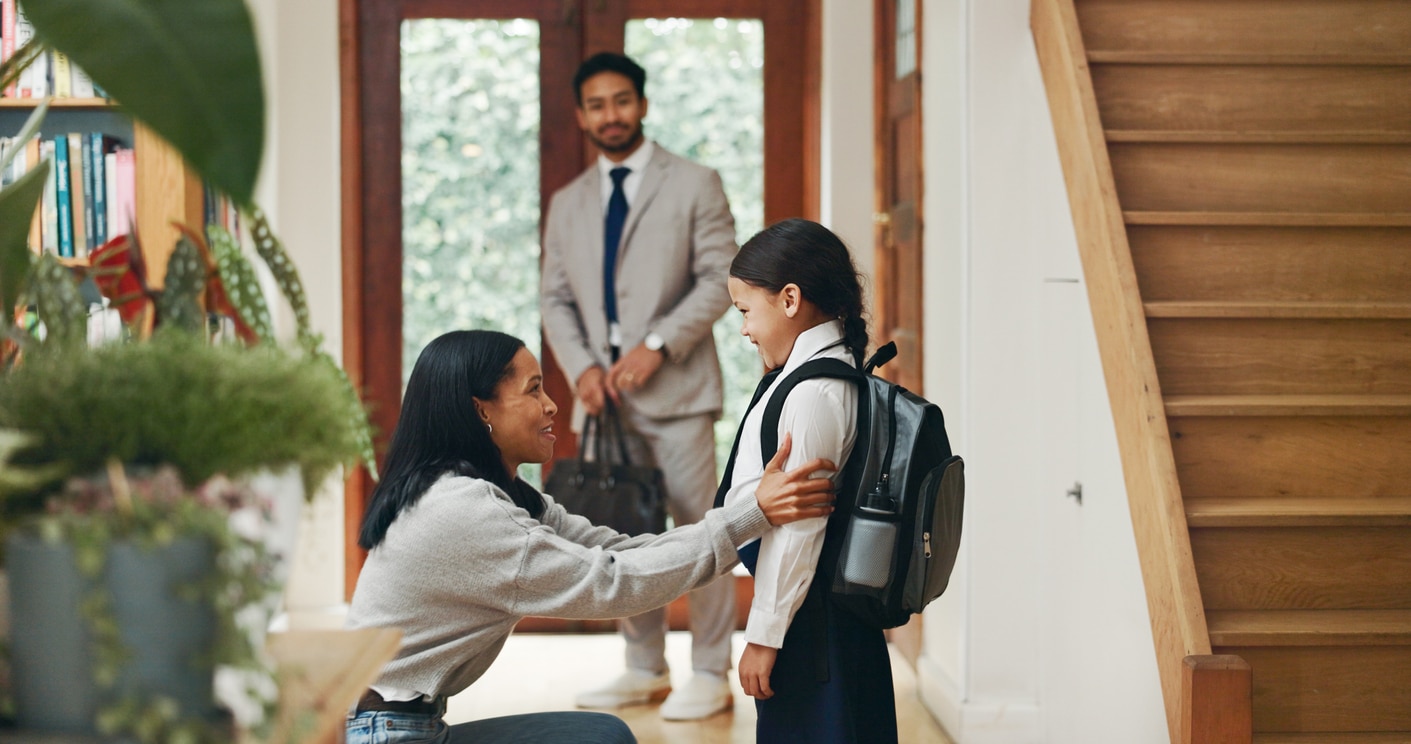 A mother adjusts her daughter's school backpack while a father in a suit watches by the door