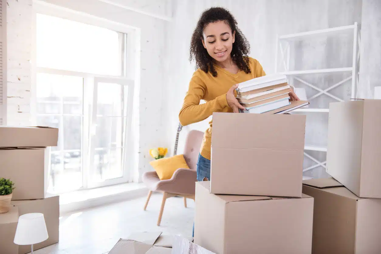 A woman in a yellow sweater smiles while packing books in preparation for moving for education