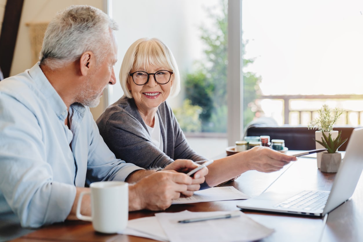 A smiling elderly couple at a table with a laptop, working together after moving for retirement