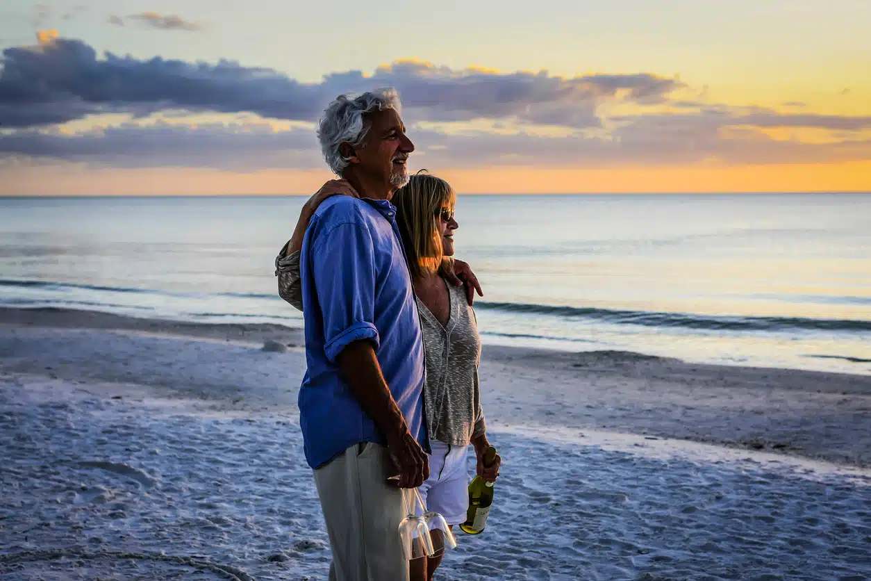 A smiling, older couple embraces on a sandy beach, watching the sunset by the ocean