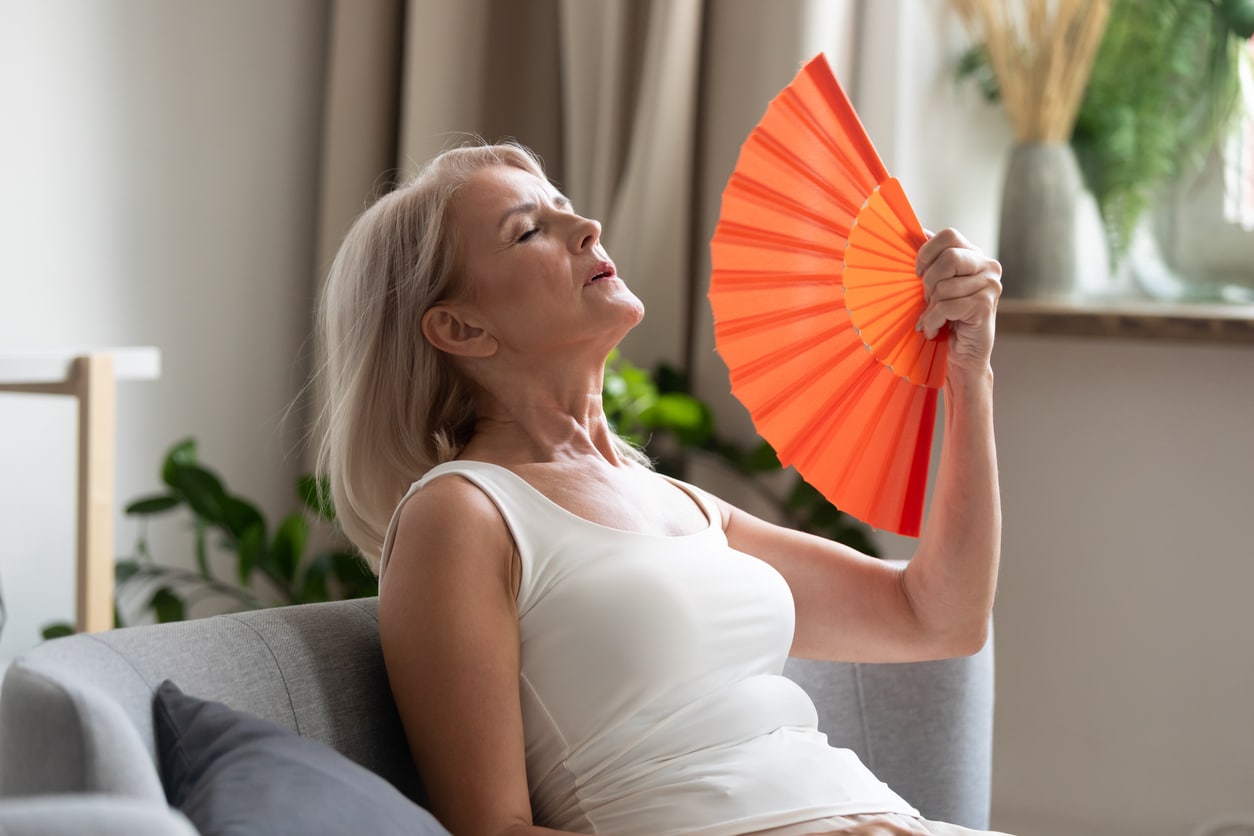 An older woman feels hot and uses an orange fan while sitting on a couch