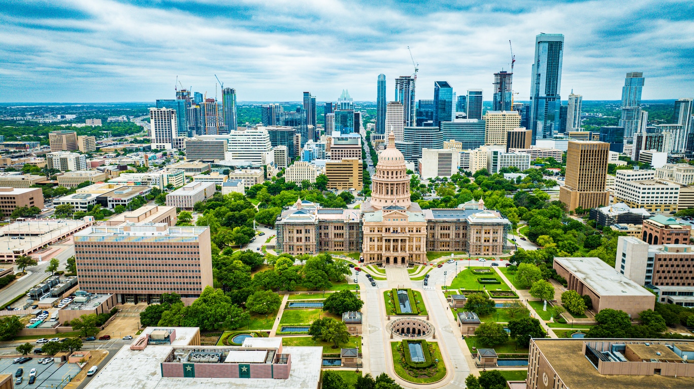 The Texas State Capitol building is centered in front of the Austin skyline under a bright blue sky