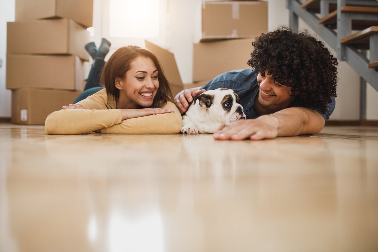 A happy couple relaxes on the floor with their bulldog, surrounded by moving boxes in their new home