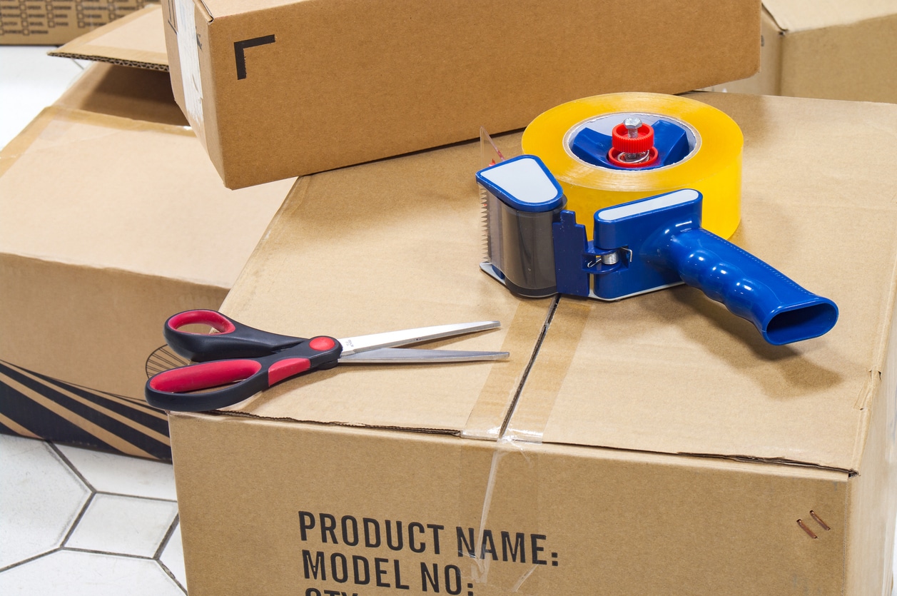 A tape dispenser with tape and scissors on top of cardboard boxes, must-haves for a packing checklist for moving