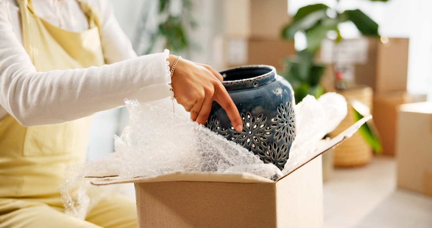 A woman puts a fragile, blue decorative pot into a moving box with bubble wrap