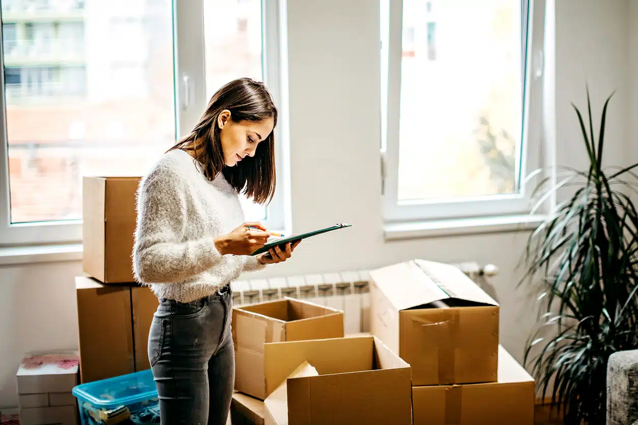 A woman in a white sweater stands among cardboard moving boxes and ticks off her packing checklist for moving