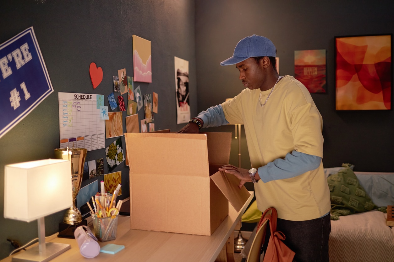 A young Black man in a blue cap opening a cardboard box at a desk in a decorated room