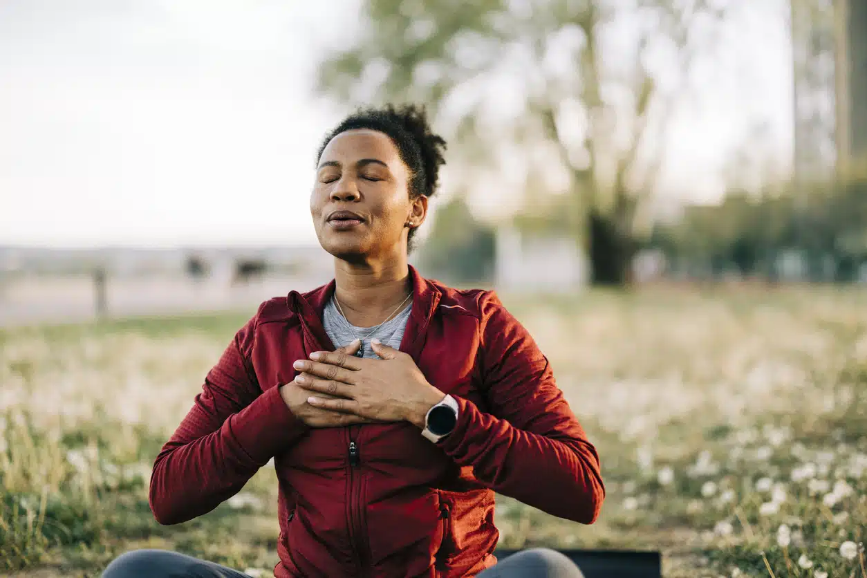 A woman in a red jacket sits in a field with her eyes closed and hands over her heart, practicing mindfulness