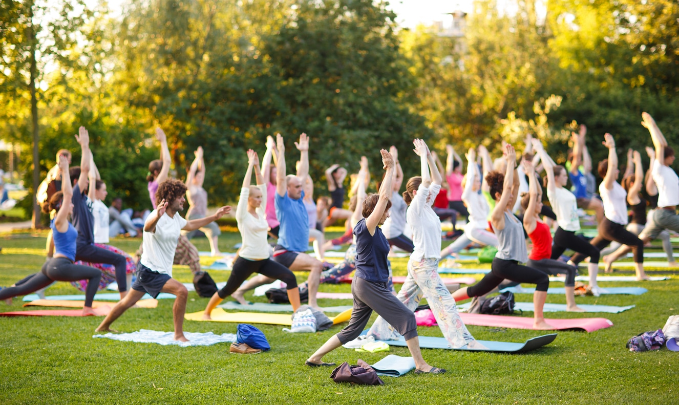 A large group of people practice yoga together in a sunlit park, performing a warrior pose on colorful mats