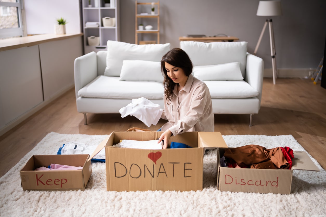 A woman sorts clothes into boxes labeled Keep, Donate, and Discard while sitting on a rug in her living room