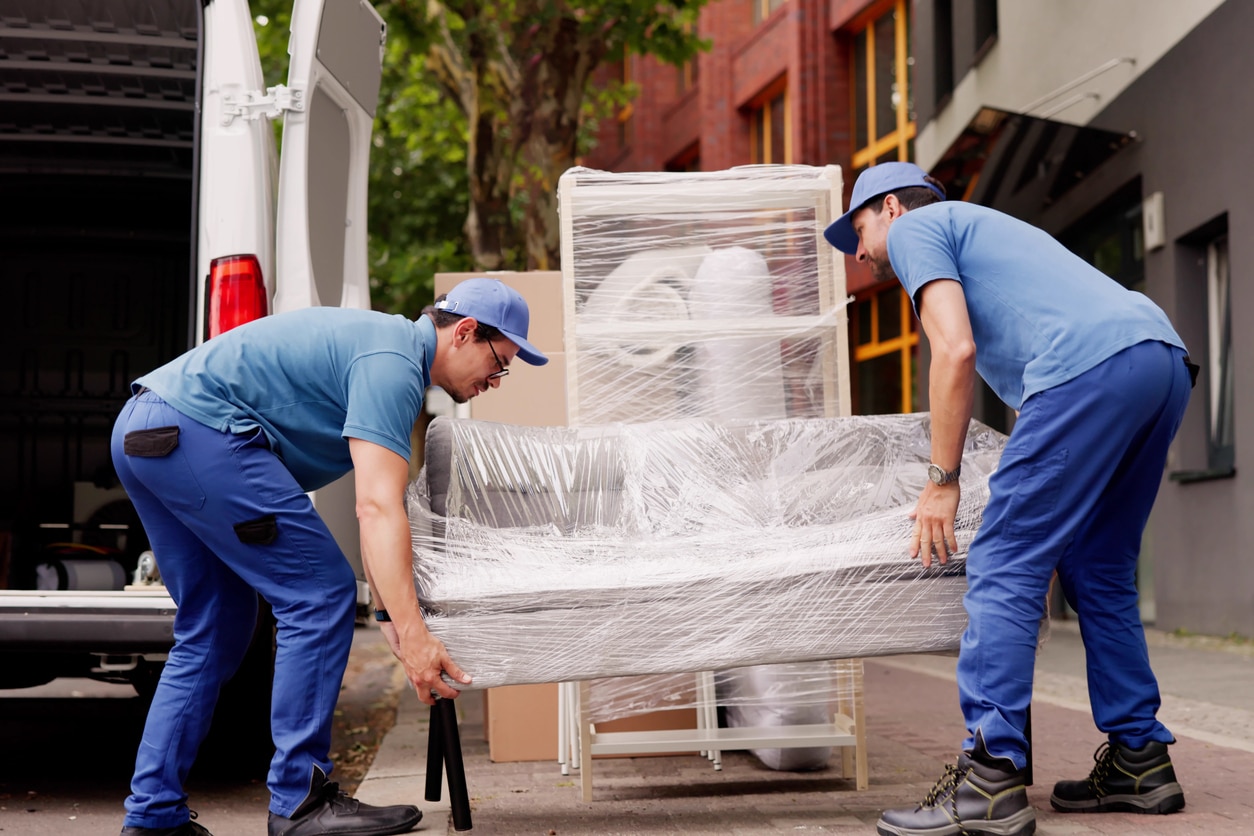Two movers in blue uniforms lift a plastic-wrapped gray sofa during a move from Atlanta to Chicago