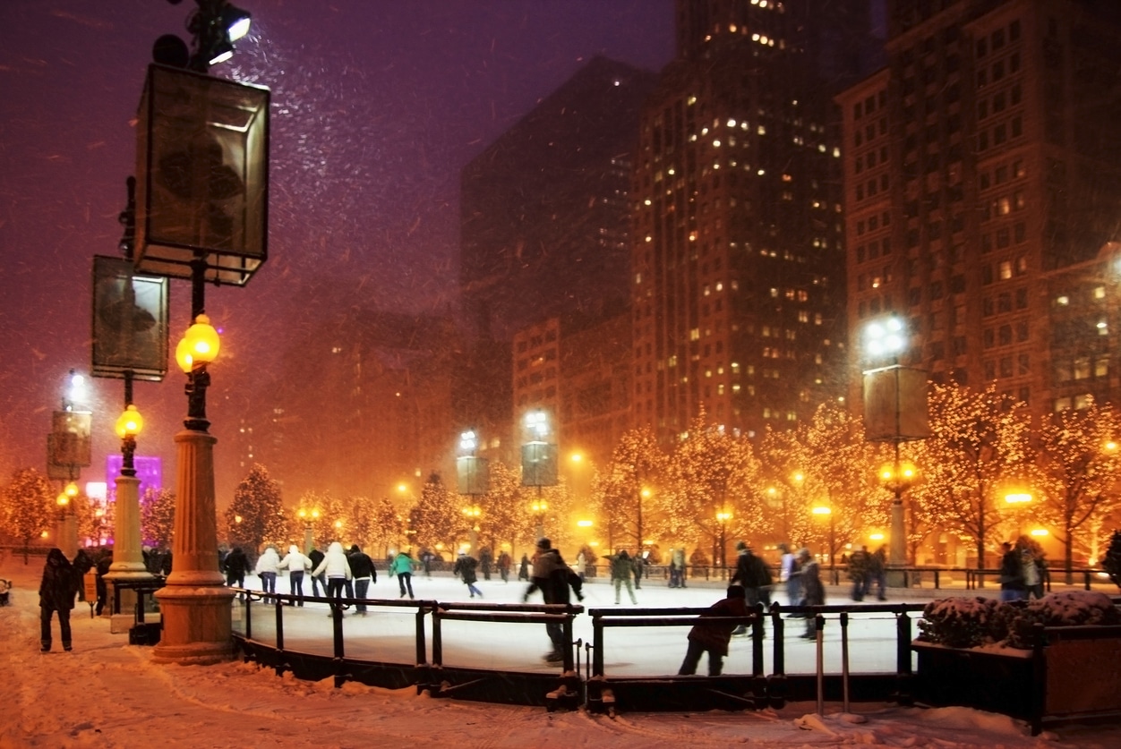 People ice skate in Chicago on an outdoor rink surrounded by lit trees and city buildings during a heavy snowfall