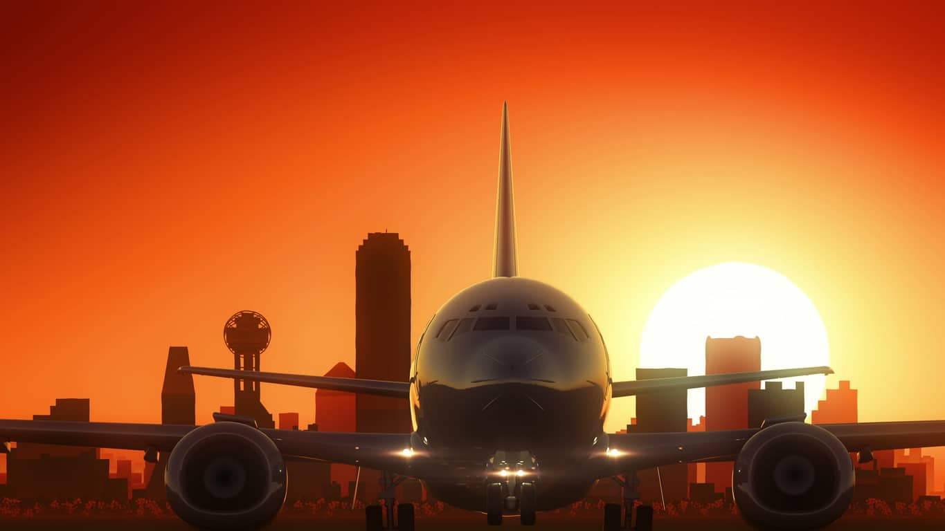 An airplane sits on a runway at sunset, with the silhouette of the Dallas skyline and Reunion Tower in the background