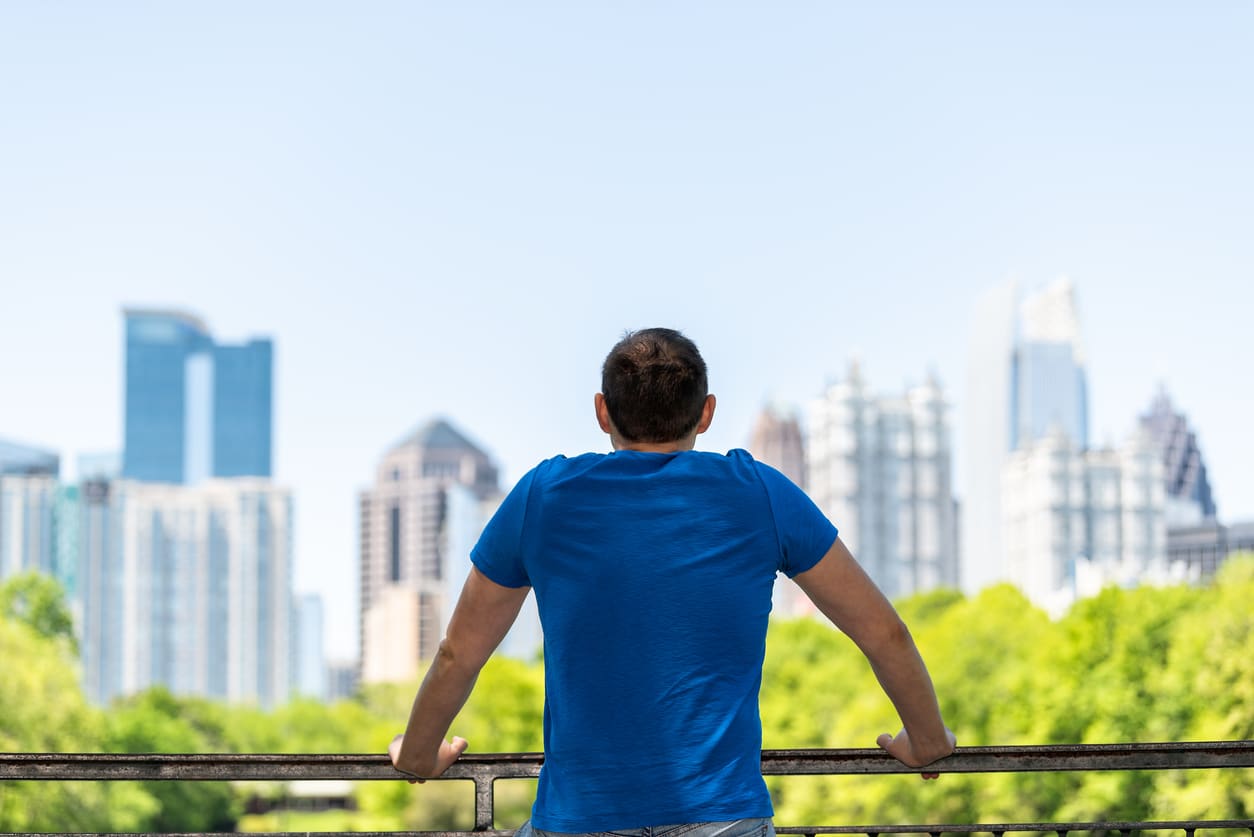 Young man standing leaning on railing in Piedmont Park in Atlanta, Georgia with scenic cityscape skyline of urban city skyscrapers downtown, Lake Clara Meer A man in a blue t-shirt stands at a railing, looking out at the Atlanta skyline after moving from Houston to Atlanta