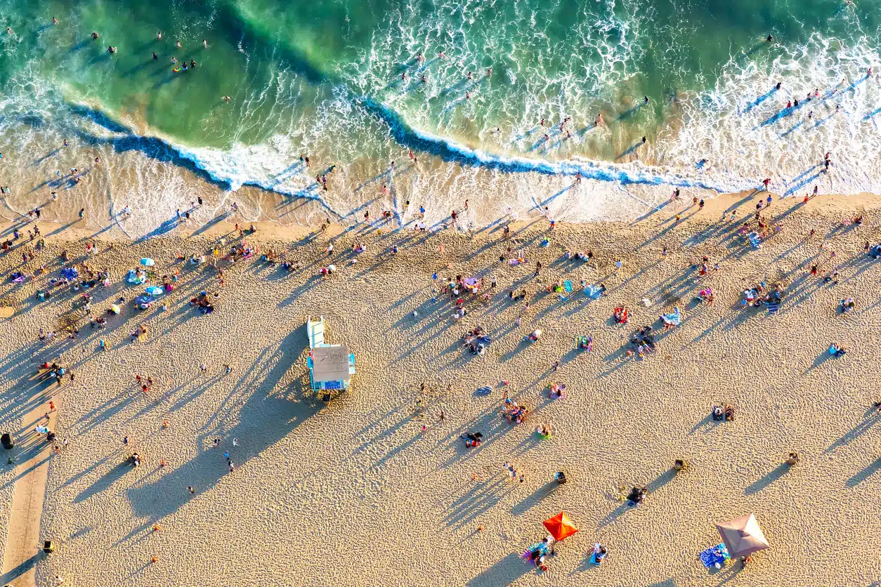 High-angle view of a crowded Los Angeles beach with turquoise waves, golden sand, and people casting long shadows
