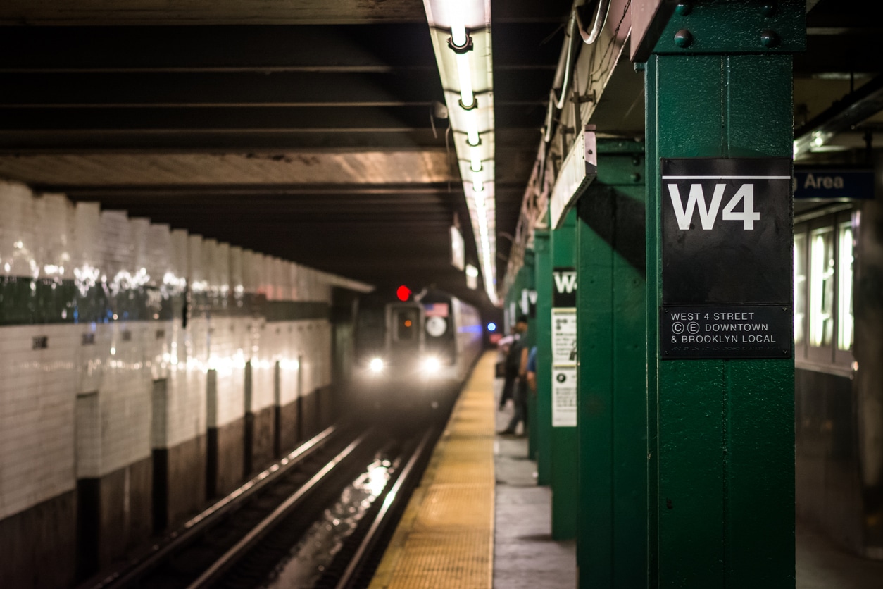 A subway train approaches the West 4 Street station platform in New York City, seen from behind a green support pillar