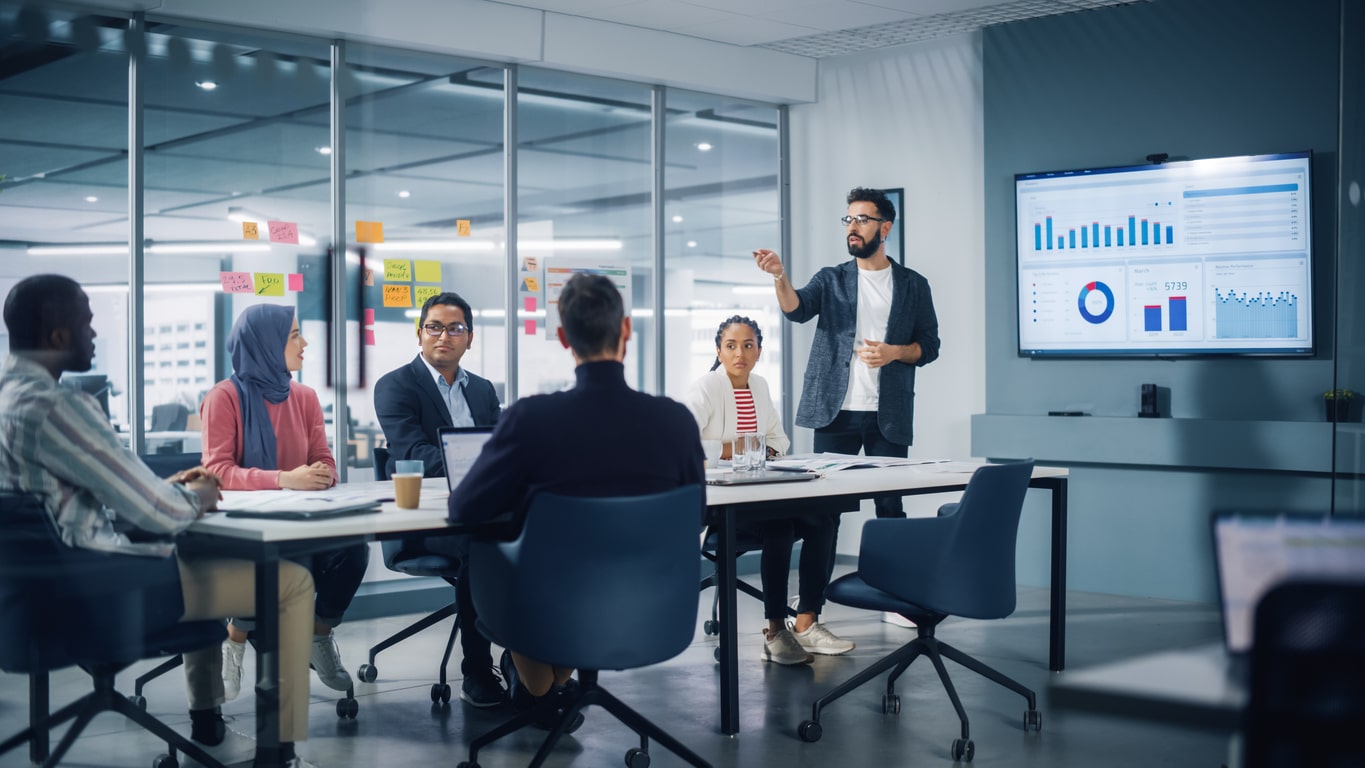 Diverse Modern Office: Businessman Leads Business Meeting with Managers, Talks, uses Presentation TV with Statistics, Infographics. Digital Entrepreneurs Work on e-Commerce Project. A man leads a business meeting after moving from Washington DC to NYC