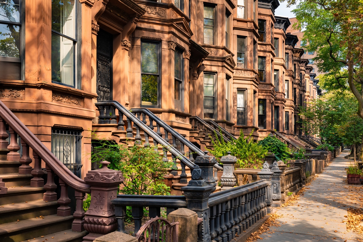 A row of classic brownstone apartment buildings with stone stoops and black railings on a sunlit, tree-lined street