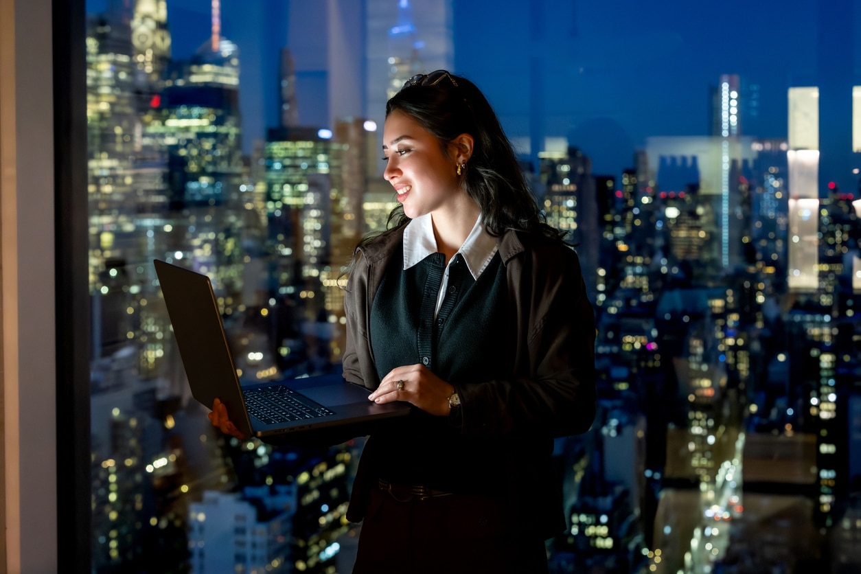 A woman smiling while using a laptop in front of a window overlooking a bright New York City skyline at night