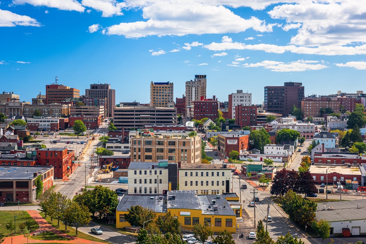 The Portland, Maine, skyline with various brick and modern buildings under a bright blue, cloud-filled sky