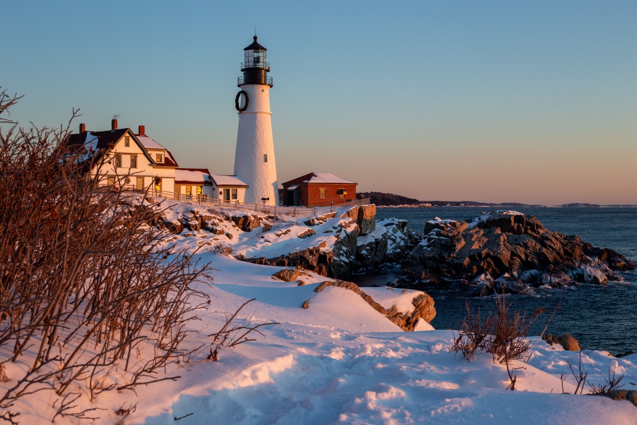A historic Portland, Maine lighthouse stands on a rocky, snow-covered coast against a pale winter sky