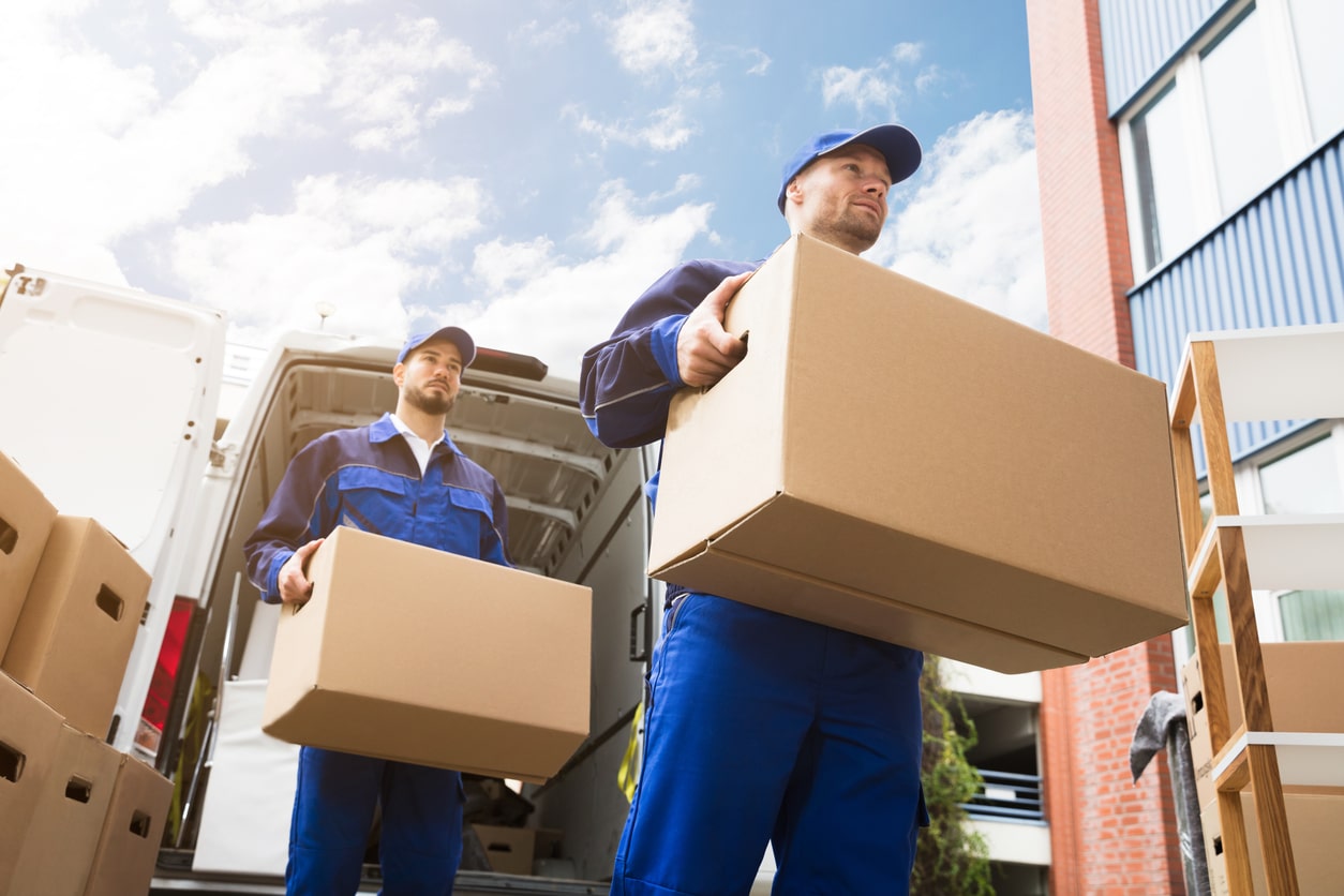 Two movers in blue uniforms carry large moving boxes away from a white moving van under a bright sky