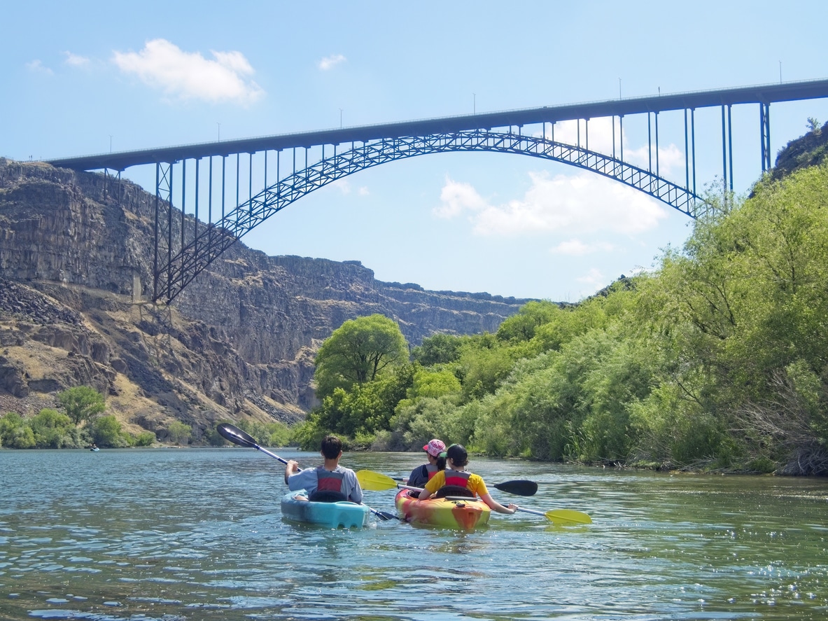 Three people kayaking down a calm Idaho river flanked by evergreen trees under a bright blue sky