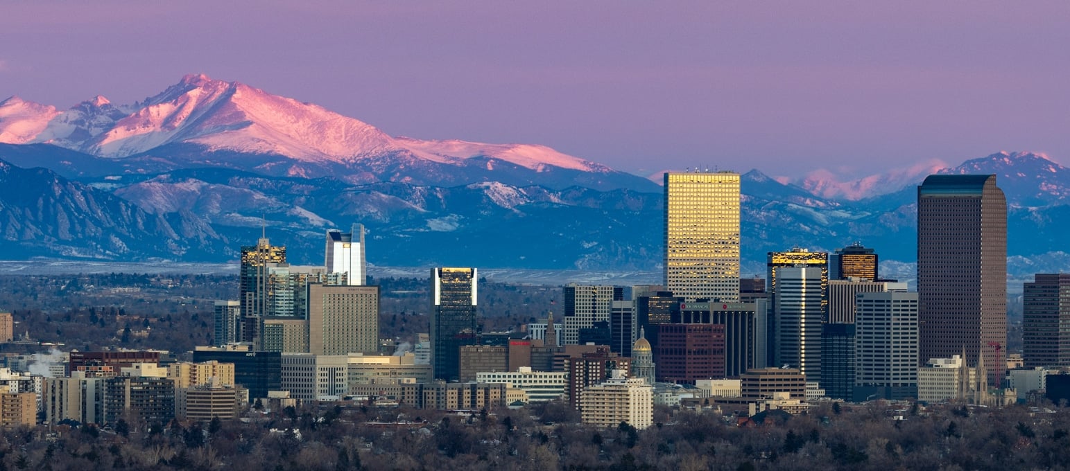 The Denver, Colorado, skyline at dusk with skyscrapers lit by the setting sun against a background of snow-capped mountains