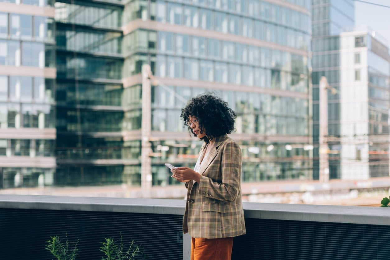 A professional woman with curly hair stands outside looking at her phone in one of the best cities for tech jobs
