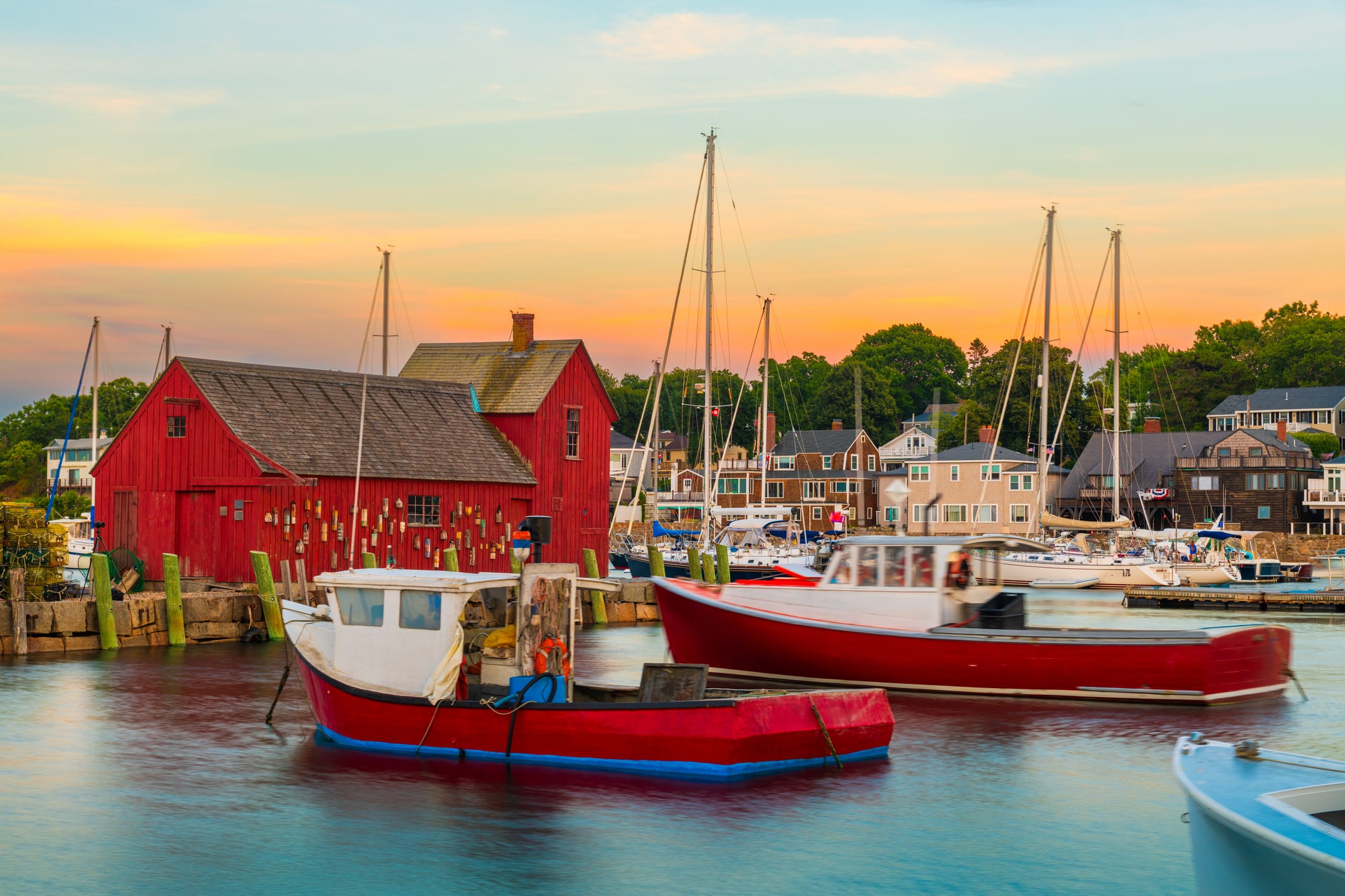 Small boats float in a New England harbor next to a historic red wooden fishing shack under a vibrant sunset sky