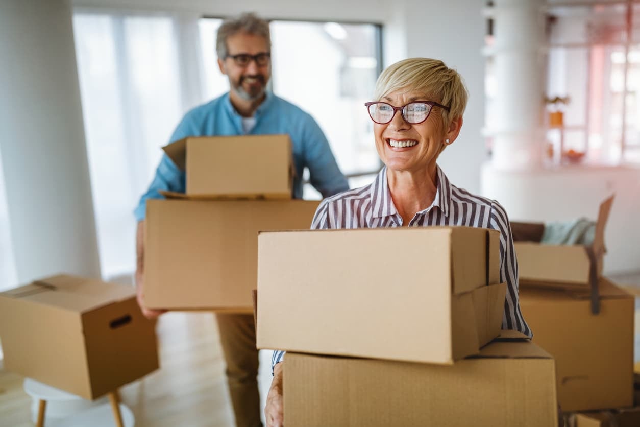 A smiling older couple carries stacks of moving boxes as they get ready to downsize their home for retirement
