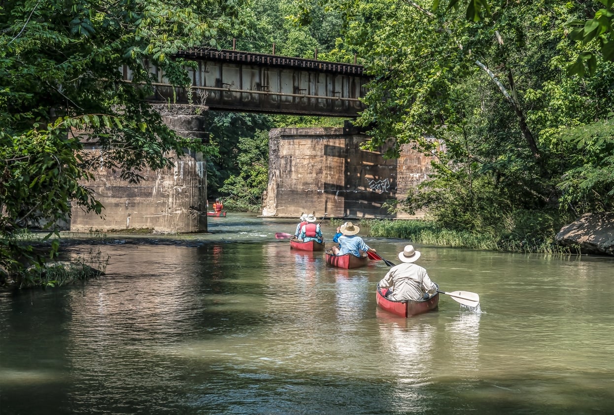 Several people paddle red canoes down a shaded Alabama river toward an old concrete and steel railroad bridge