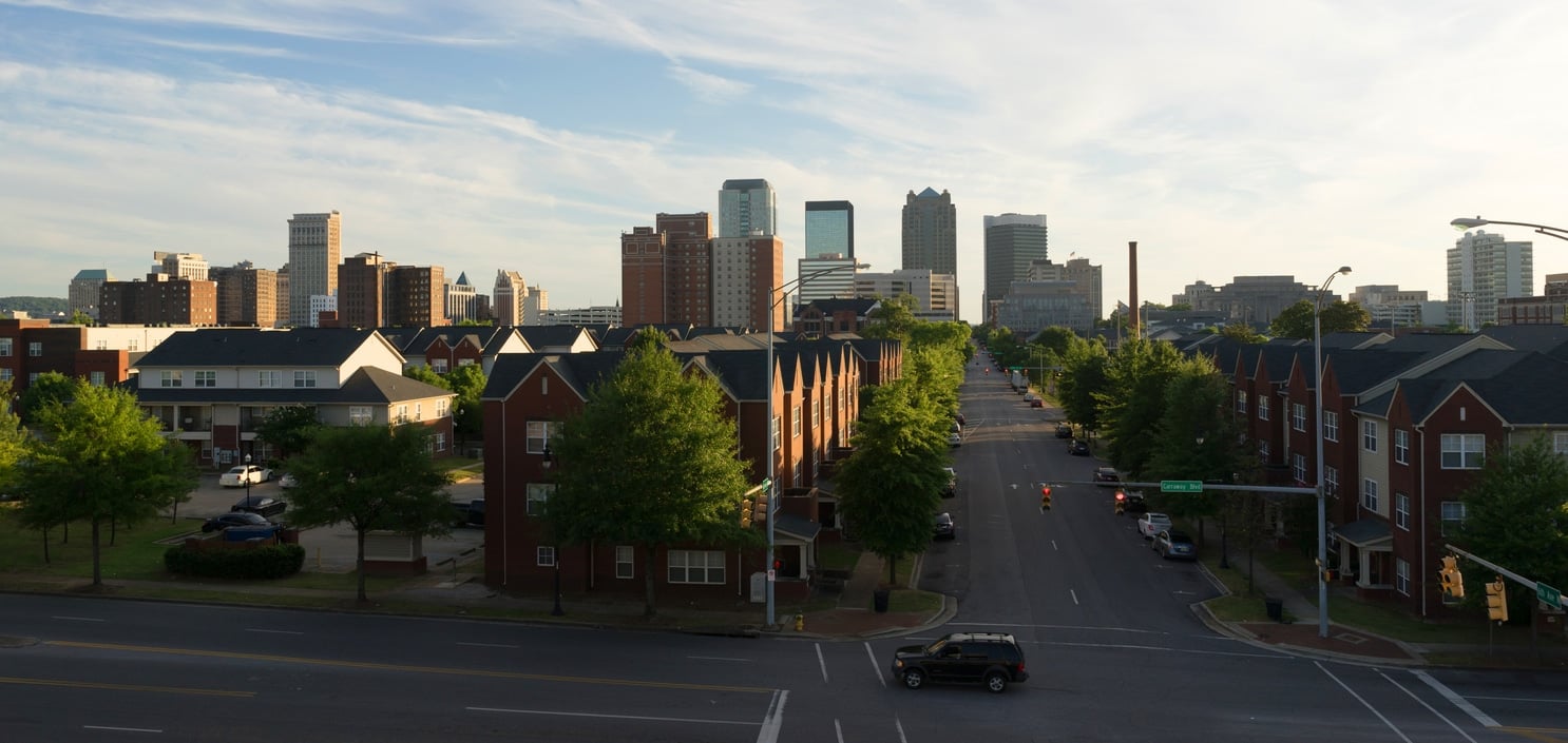 A dusky Alabama street lined with brick apartment buildings leading toward a distant downtown skyline