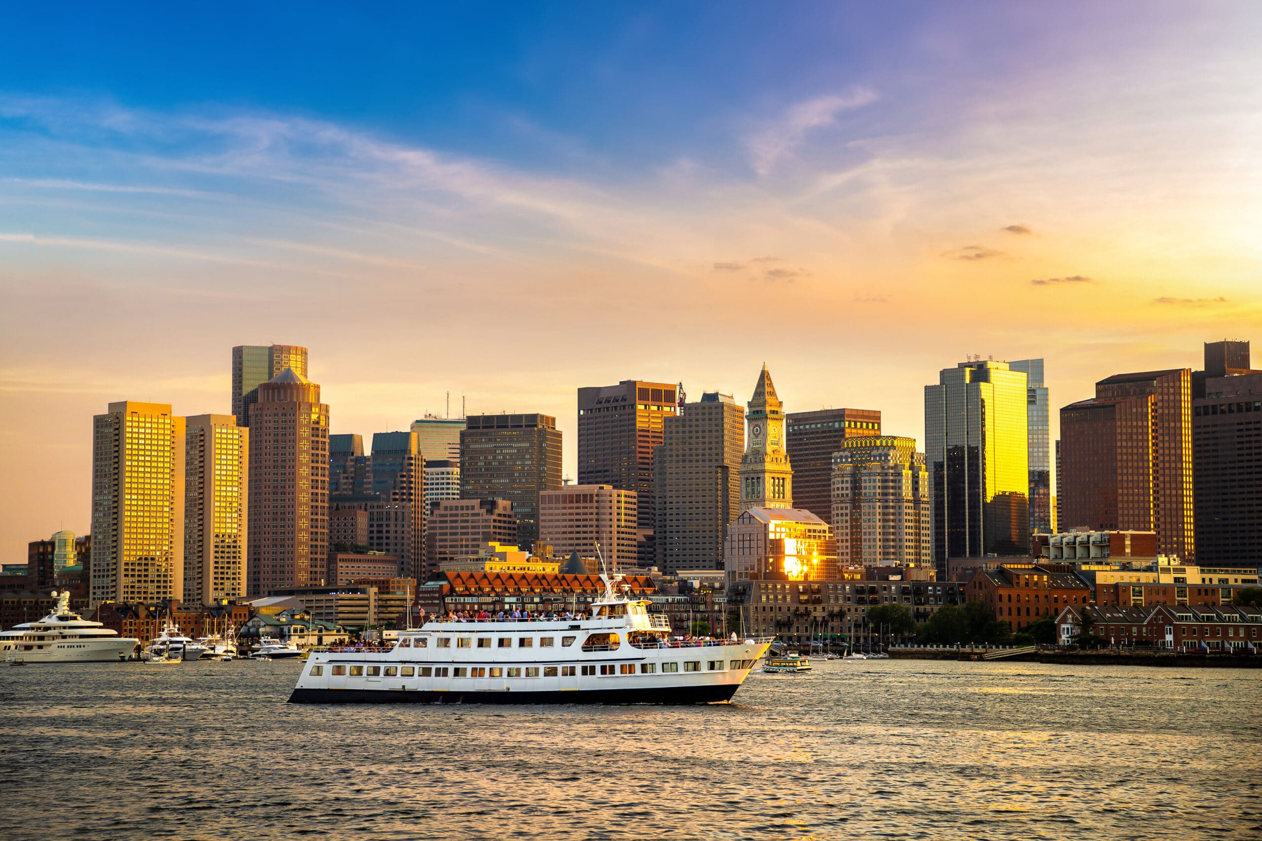 A large white ferry boat cruises across open water toward a sunlit Boston skyline during a golden sunset