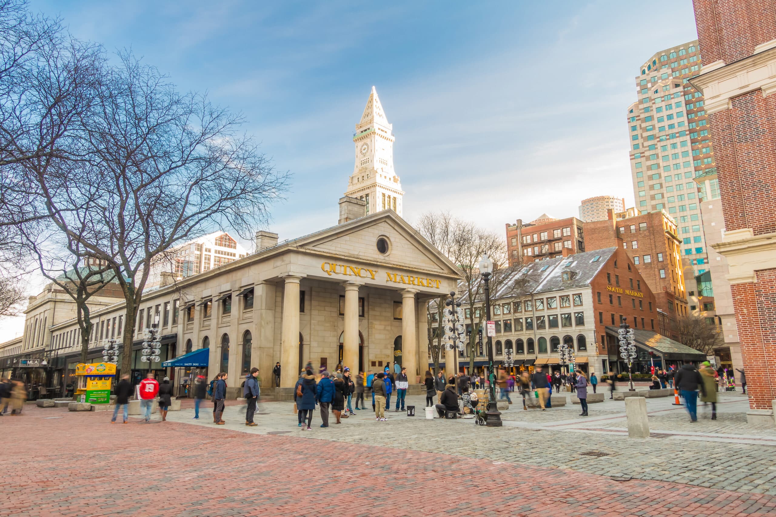People walk outside a historic stone market building with a columned entrance under a clear blue sky in NYC