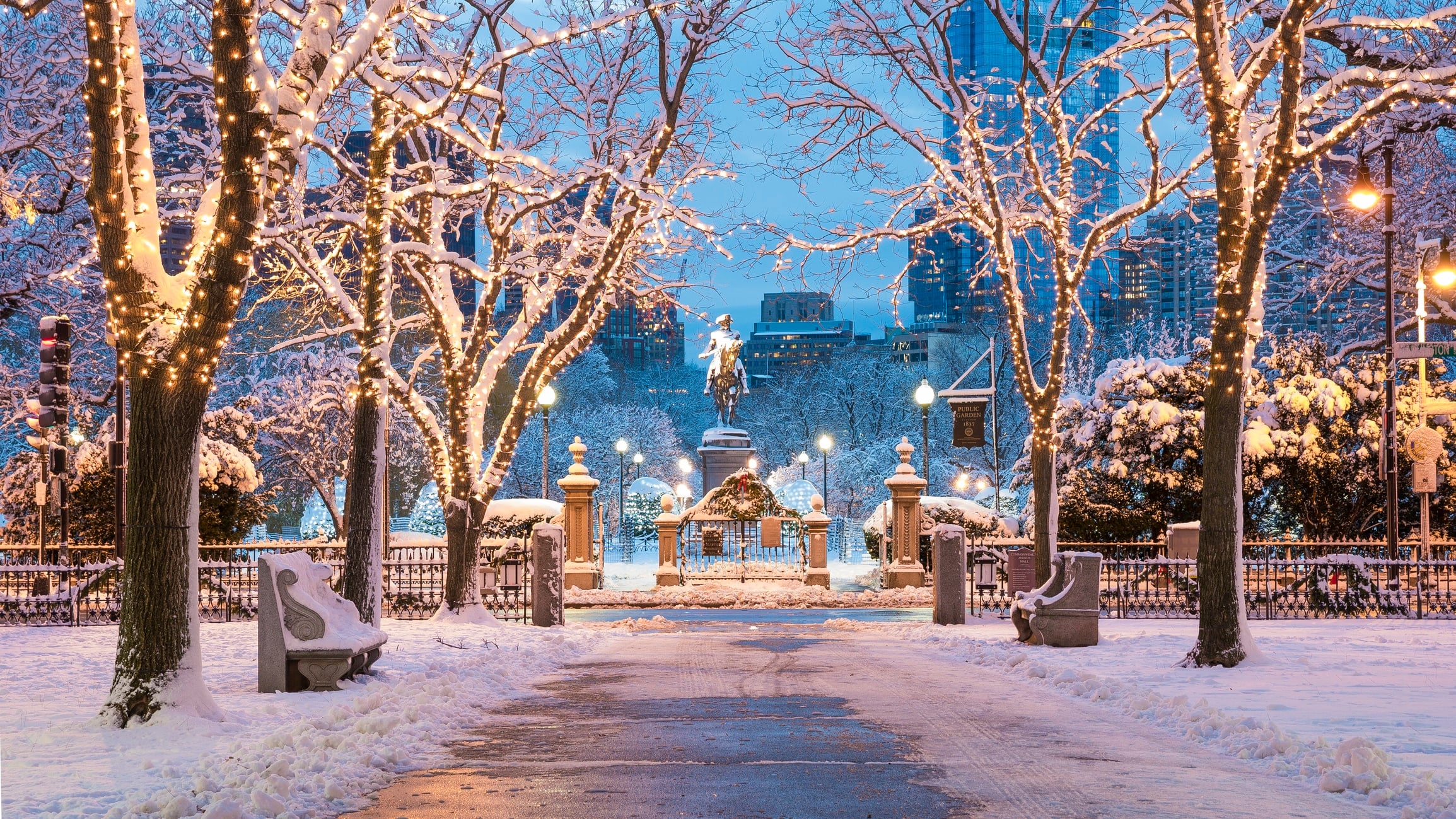 A snow-covered NYC park entrance at twilight with trees wrapped in warm lights