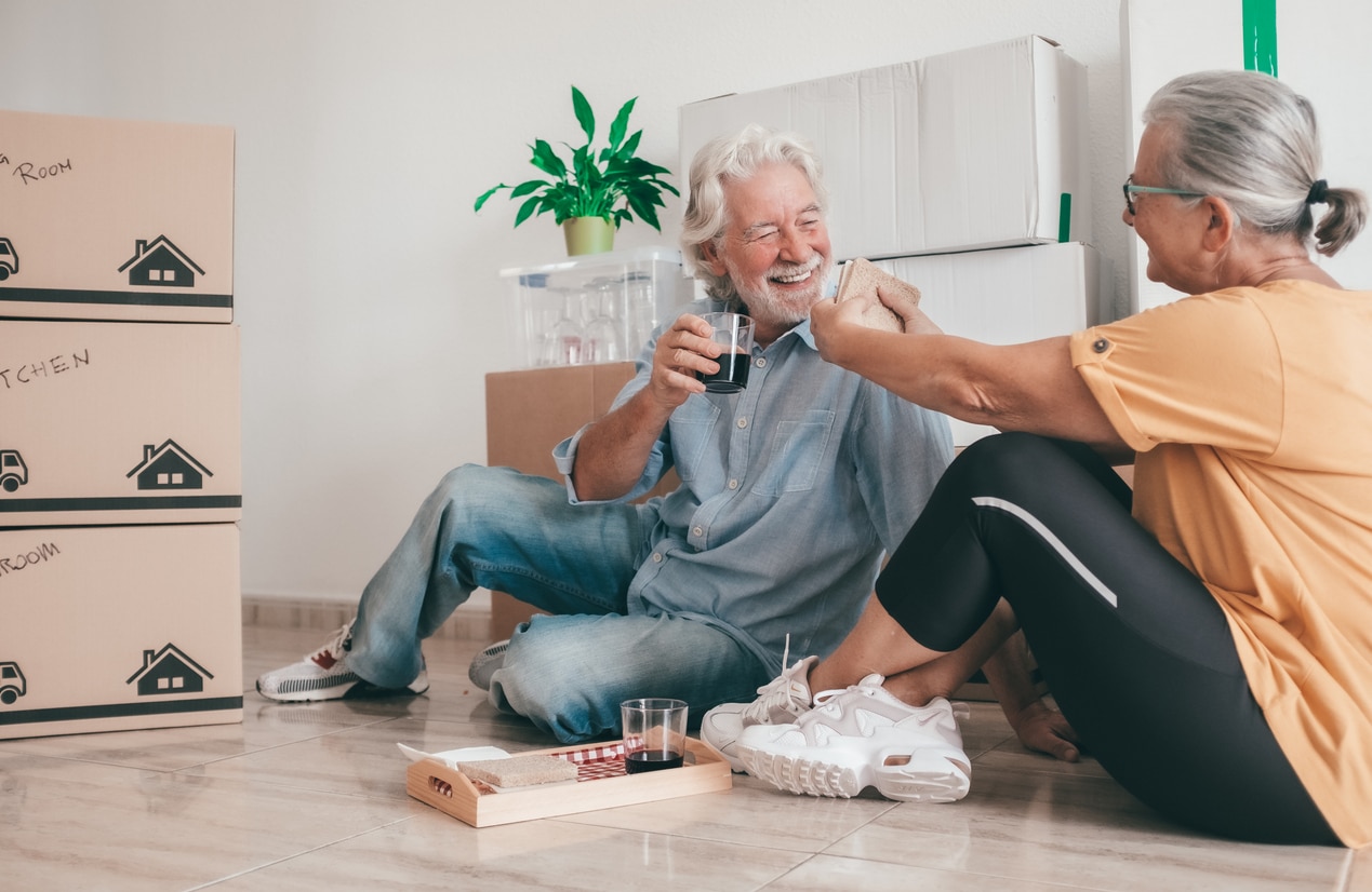 Cheerful senior couple, happily moving into their new apartment, takes a break sitting on the floor surrounded by moving boxes. They enjoy food and drinks sharing a joyful and relaxed moment together A smiling senior couple takes a break on the floor, sharing snacks and drinks surrounded by stacked moving boxes