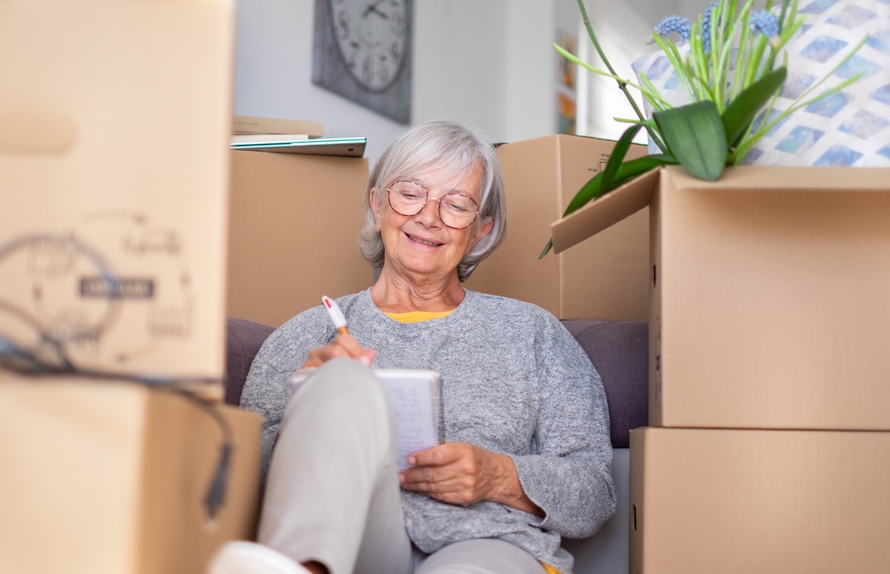 Happy senior woman involved in moving house sitting among cardboard boxes making note of purchases and to-dos, concept of moving, retirement, new life, buying, renting, apartment, house A smiling senior woman sits on the floor writing in a notebook, surrounded by moving boxes