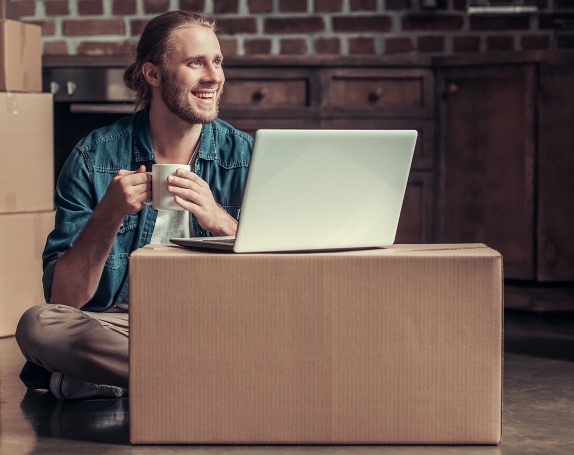 A smiling man sits on the floor holding a mug next to a moving box as he plans moving to another state without a job
