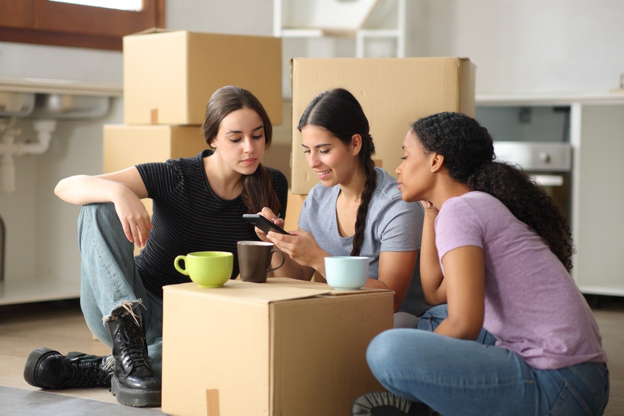 Three young women sit on the floor looking at a phone, surrounded by moving boxes