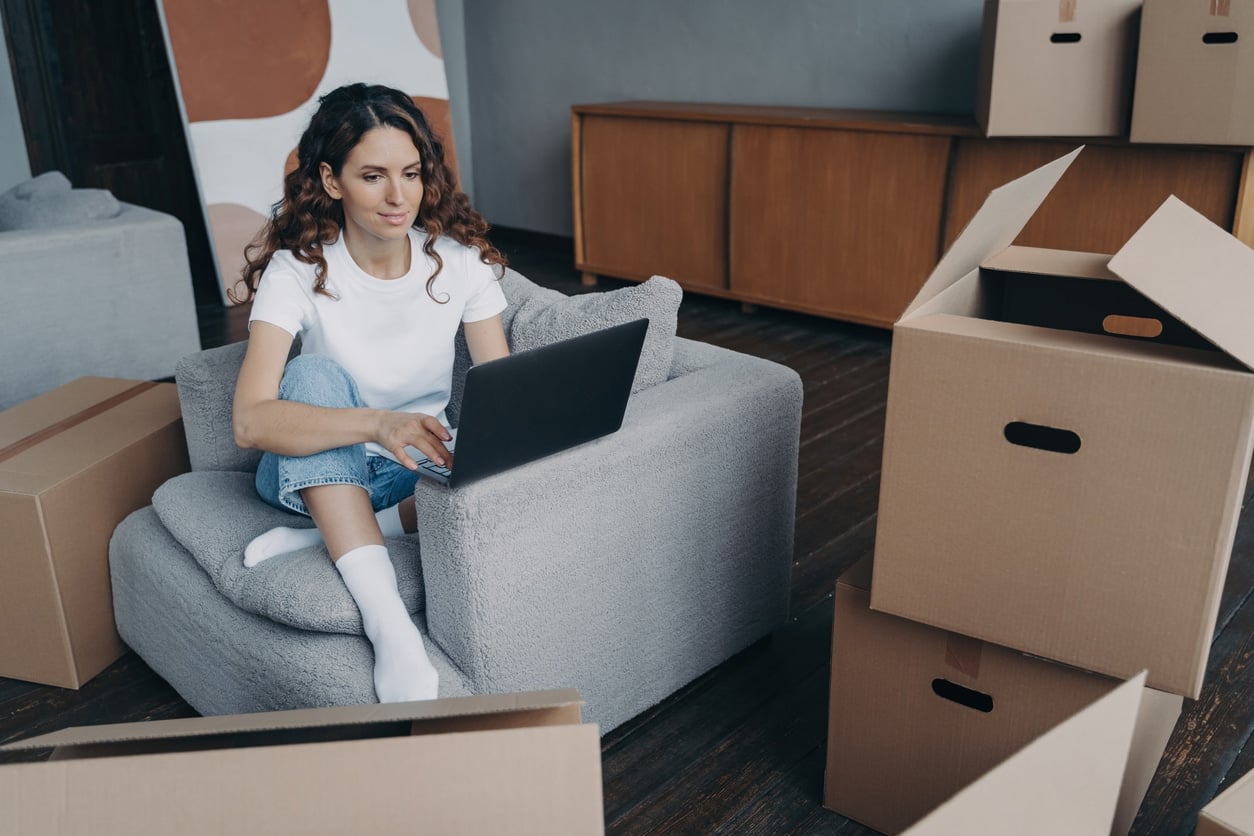 A woman sits in an armchair using her laptop surrounded by moving boxes, planning how to move without a job
