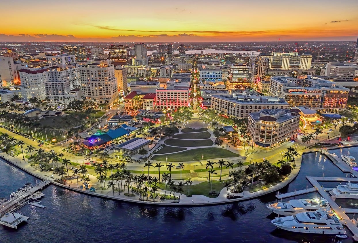 An aerial view of West Palm Beach neighborhoods illuminated at dusk, in front of a waterfront park and a marina